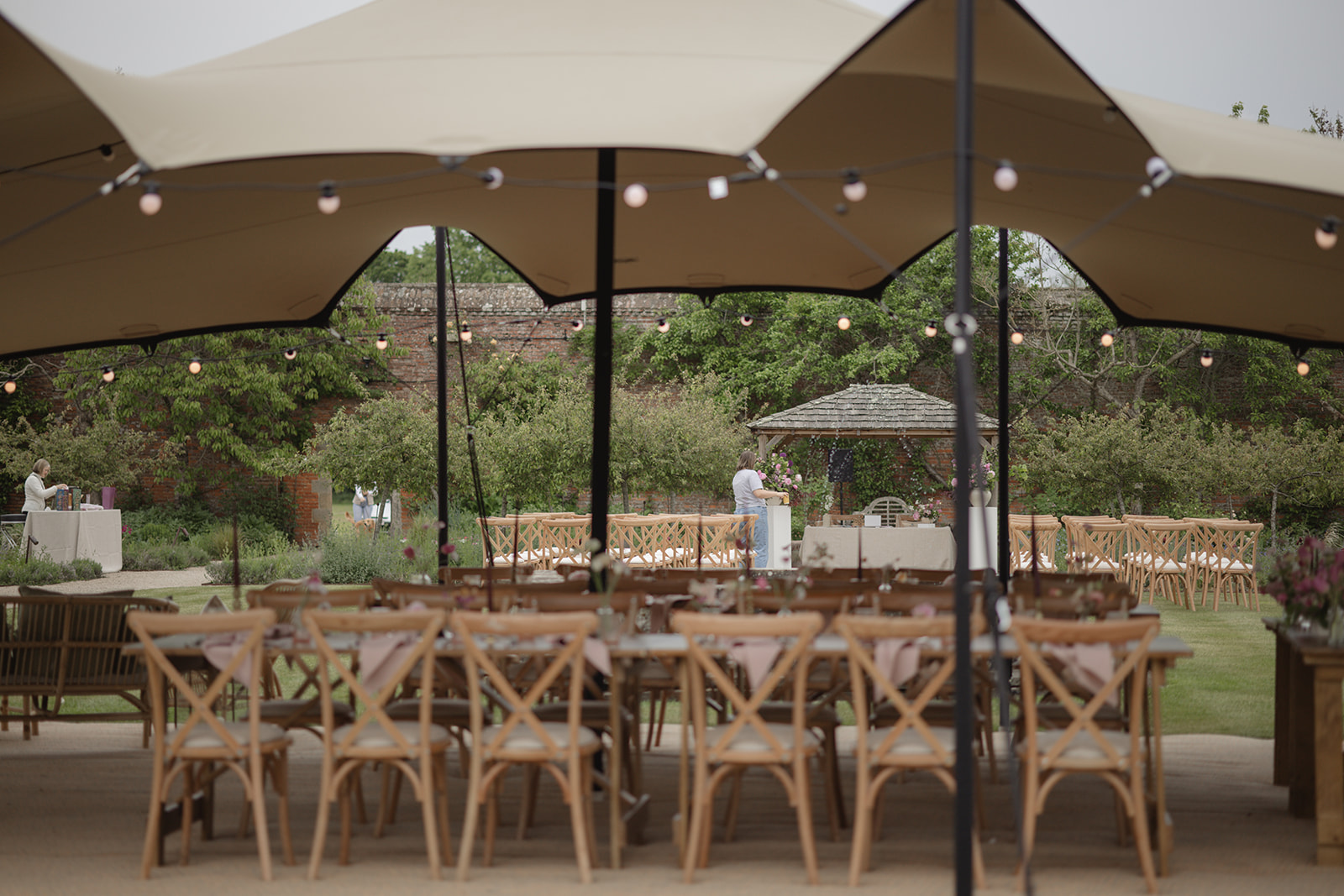 Rustic tables and cross-back chairs set for a wedding under a stretch tent at The Walled Garden, Cowdray
